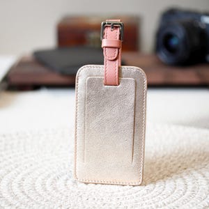 May include: A rectangular luggage tag with a metallic gold finish. It features a pink leather strap and buckle at the top. The tag has a textured surface and a smaller rectangular section on the front.