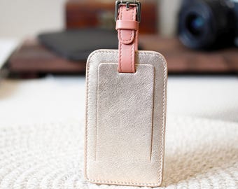 May include: A rectangular luggage tag with a metallic gold finish. It features a pink leather strap and buckle at the top. The tag has a textured surface and a smaller rectangular section on the front.