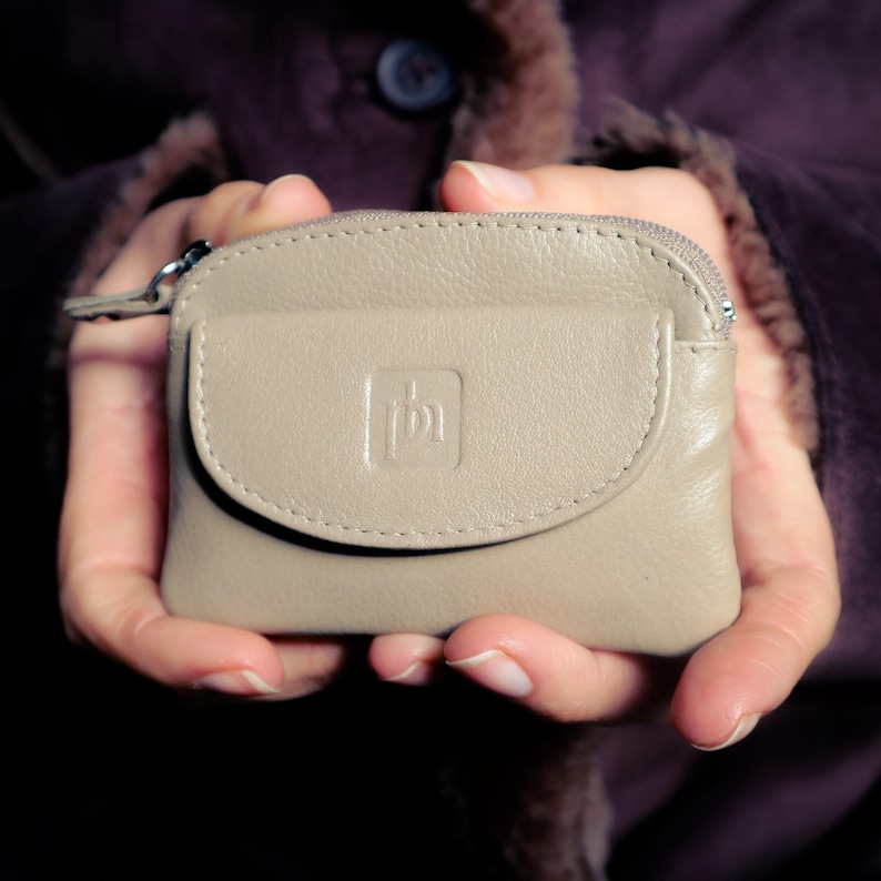 May include: A small, beige leather coin purse featuring a top zipper closure and a front flap pocket with a debossed square logo. The purse has a smooth, rounded rectangular shape with visible stitching along the edges of the flap and body.