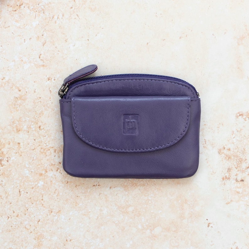 May include: A small purple leather coin purse featuring a top zipper closure and a front flap pocket with a debossed square logo. The rectangular pouch has rounded corners and a smooth finish, providing a compact storage solution for small items.