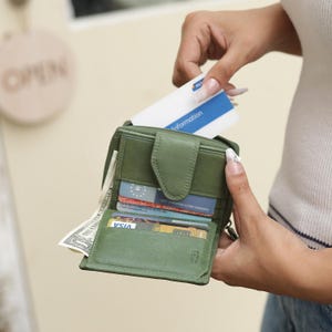 May include: An olive green leather bifold wallet featuring a snap button closure, multiple interior card slots, and a dedicated compartment for paper currency. The wallet is open, displaying several credit cards and a single US dollar bill inside.