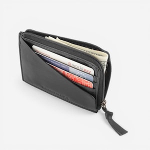 Op de afbeelding: Zwart leren portemonnee met ritssluiting, gedeeltelijk open om contant geld en creditcards te laten zien. De portemonnee heeft een minimalistisch ontwerp en een klein, bevestigd treklipje. Zichtbare kaarten zijn een lidmaatschapskaart en een Worldpay-kaart.