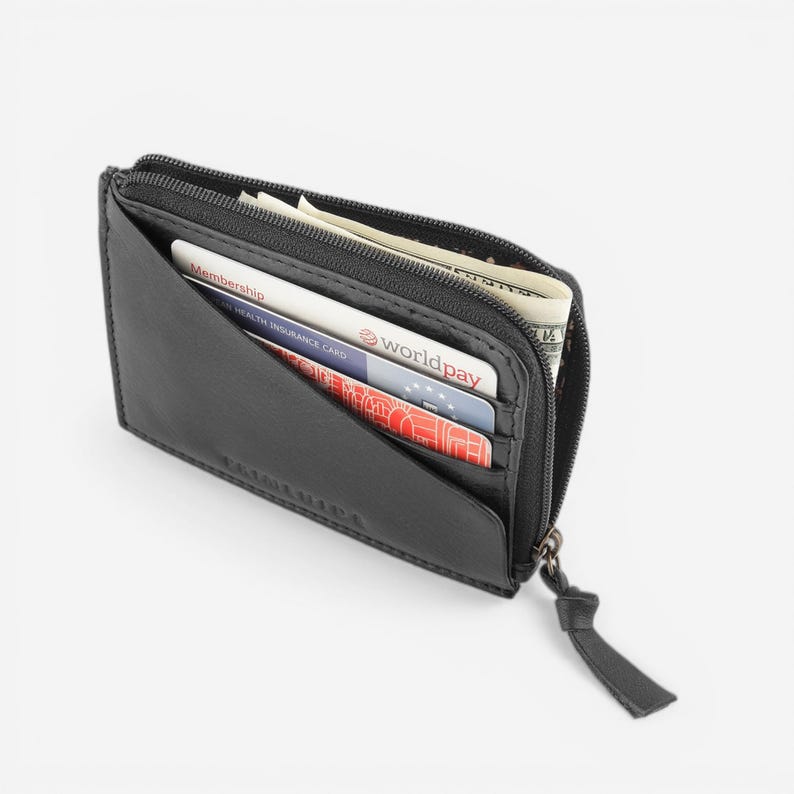 Op de afbeelding: Zwart leren portemonnee met ritssluiting, gedeeltelijk open om contant geld en creditcards te laten zien. De portemonnee heeft een minimalistisch ontwerp en een klein, bevestigd treklipje. Zichtbare kaarten zijn een lidmaatschapskaart en een Worldpay-kaart.