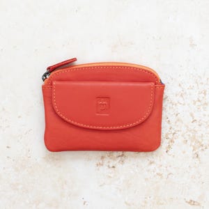 May include: A vibrant orange leather coin purse featuring a top zipper closure and a front flap pocket with a snap button. The front flap is embossed with a small square logo containing the letters ph. The purse has a smooth, matte finish.
