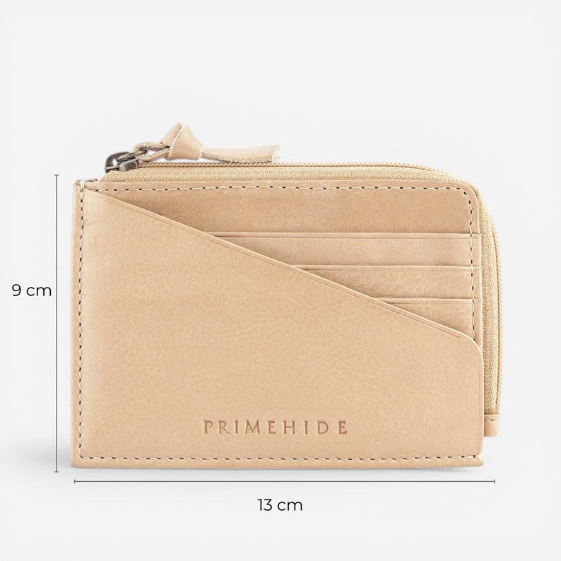 Op de afbeelding: Een beige leren portemonnee met ritssluiting en meerdere kaartsleuven. De portemonnee is 13 cm breed en 9 cm hoog. De merknaam "PRIMEHIDE" is op de voorkant in reli&euml;f aangebracht. De portemonnee is gemaakt van glad, getextureerd leer.