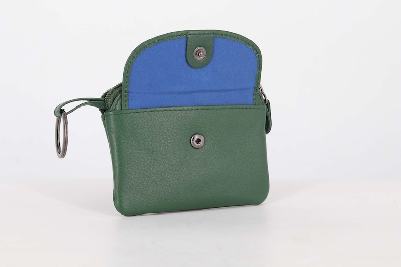May include: A forest green leather coin purse with a snap button closure and a vibrant blue interior lining. The accessory features a metal key ring attached to the side zipper pull, providing a functional design for carrying small essentials.