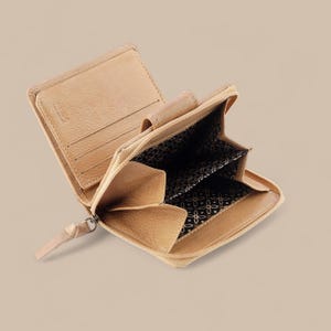 May include: A tan leather bifold wallet with a zipper closure, featuring multiple interior card slots, a coin compartment, and a black fabric lining with a repeating white geometric floral pattern. The wallet includes an RFID protection stamp.