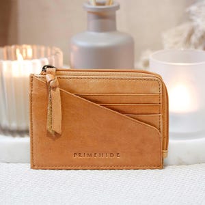 A tan leather wallet with a zip closure and several card slots. The wallet features a diagonal design and a small tassel. The brand name "PRIMEHIDE" is embossed on the front.