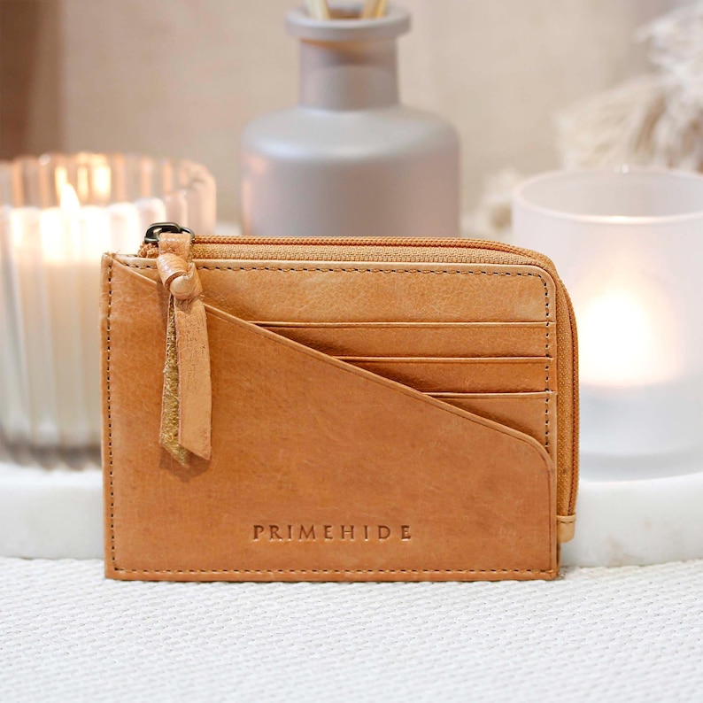 A tan leather wallet with a zip closure and several card slots. The wallet features a diagonal design and a small tassel. The brand name "PRIMEHIDE" is embossed on the front.
