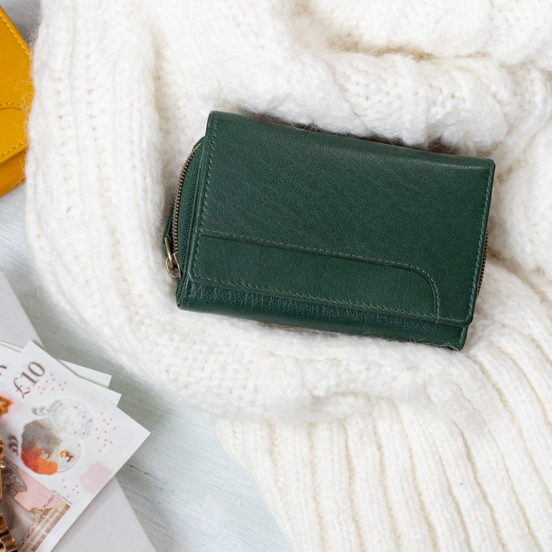 Soft leather purse in green placed on a white knit fabric with British currency notes and a watch visible nearby. This ladies wallet is an excellent unique gift for her or a gift for wife.