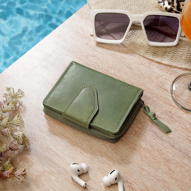 May include: A sage green leather wallet featuring a textured surface, a snap-button closure flap, and a side zipper with a matching leather pull tab. The wallet is rectangular with rounded corners and a compact, folded design.