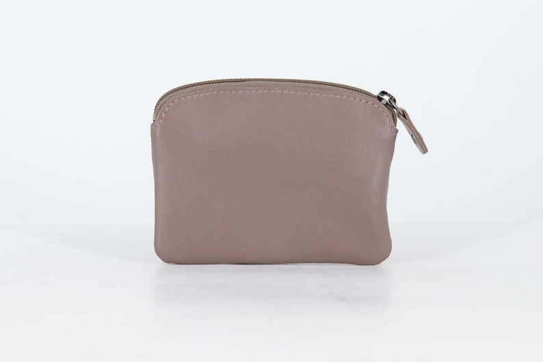 May include: A taupe-colored coin purse with a smooth, matte finish and a rounded top edge. The pouch features a matching taupe zipper closure along the top, complete with a small, thin leather pull tab attached to the silver-toned metal zipper slider.