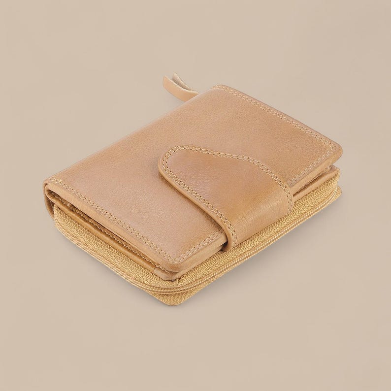May include: A light tan leather wallet featuring a wrap-around zipper closure and a front flap secured with a snap button. The wallet has visible stitching along the edges and a smooth, matte finish, showcasing a compact and functional design.