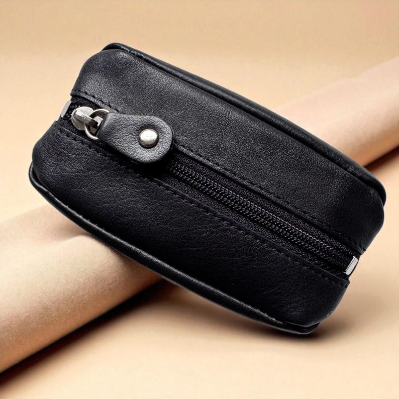 A black leather key case with a zip closure and a small, silver-coloured metal pull. The case is rectangular with rounded edges and a small, circular detail near the zip pull. The case is designed to hold keys and coins.