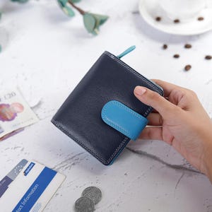 May include: A bi-fold wallet crafted from dark navy blue leather with a contrasting bright blue snap-button closure tab. The wallet features visible white stitching along the edges and a small matching blue zipper pull on the side for access.