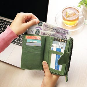 May include: A green leather bi-fold wallet, open to display credit cards and several Bank of England &pound;20 notes. The wallet has a zip closure and multiple compartments. A cup of tea is in the background.