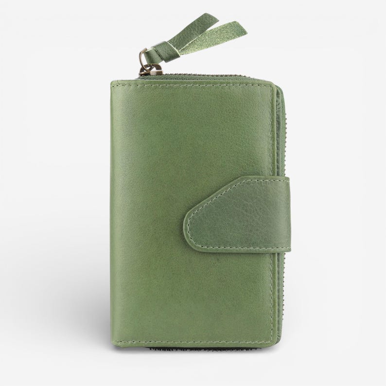 May include: An olive green leather wallet with a zip and snap closure. The wallet has a small leather pull tab attached to the zip and a decorative leather accent on the top. The wallet is approximately 12 cm high.