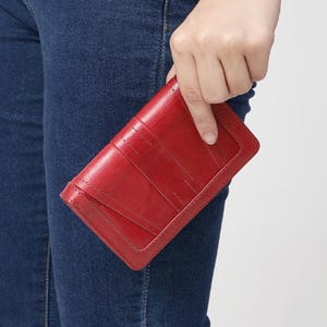 May include: A rectangular red leather wallet featuring a smooth, polished finish with visible stitched detailing along the edges and a structured flap closure. The wallet displays a deep crimson hue and a clean, minimalist design with precise seams.