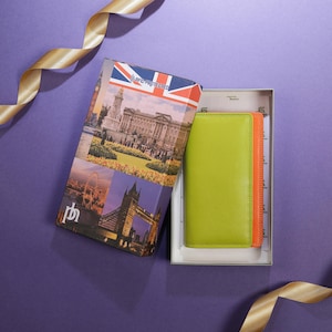May include: A green and orange leather wallet inside a gift box. The box features images of London landmarks and a Union Jack flag. The box is open, revealing the wallet. A gold ribbon is in the background.