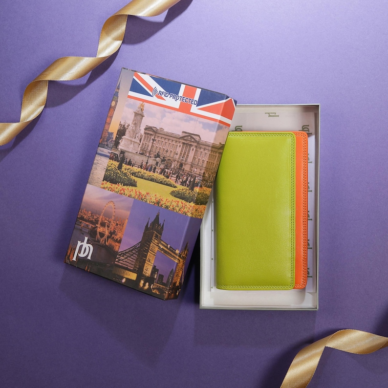 May include: A green and orange leather wallet inside a gift box. The box features images of London landmarks and a Union Jack flag. The box is open, revealing the wallet. A gold ribbon is in the background.