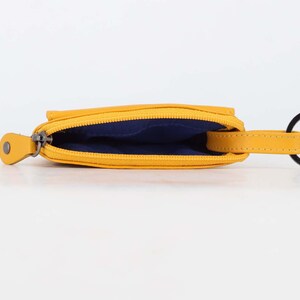 May include: A mustard yellow coin pouch with a zipper closure, featuring a dark blue interior lining and a matching yellow leather tab attached to a black circular metal key ring. The pouch is shown from an open top-down perspective.