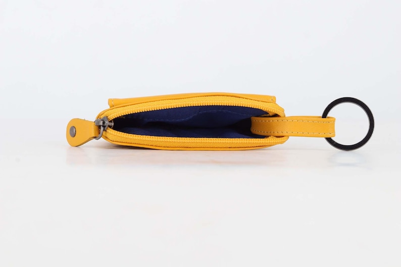 May include: A mustard yellow coin pouch with a zipper closure, featuring a dark blue interior lining and a matching yellow leather tab attached to a black circular metal key ring. The pouch is shown from an open top-down perspective.