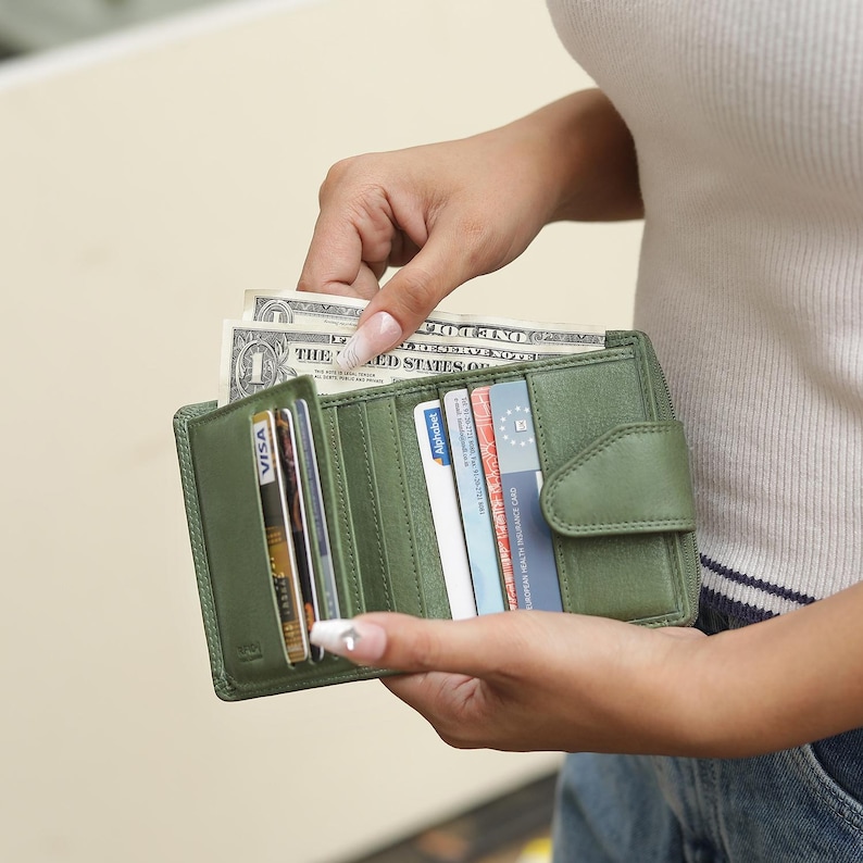 May include: A sage green leather bifold wallet featuring multiple interior card slots, a dedicated compartment for paper currency, and a snap-button closure. The wallet is open, displaying several credit cards and a one-dollar bill tucked inside.