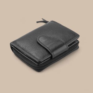May include: A black bi-fold wallet with a textured, pebbled leather surface. The wallet features a central snap-button closure flap and a wrap-around zipper that secures the main compartment. The design includes a small zipper pull tab at the top.