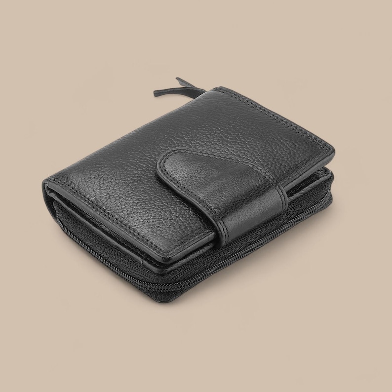May include: A black bi-fold wallet with a textured, pebbled leather surface. The wallet features a central snap-button closure flap and a wrap-around zipper that secures the main compartment. The design includes a small zipper pull tab at the top.