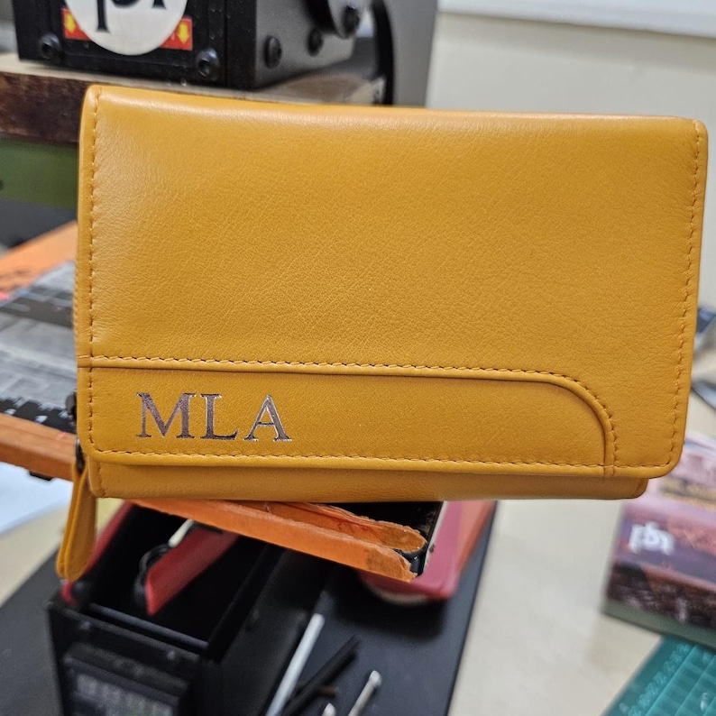 May include: A mustard yellow leather wallet featuring a horizontal stitched seam across the front flap. The letters MLA are embossed in a metallic silver font on the lower left corner of the flap. The wallet has a smooth texture and a rectangular shape.