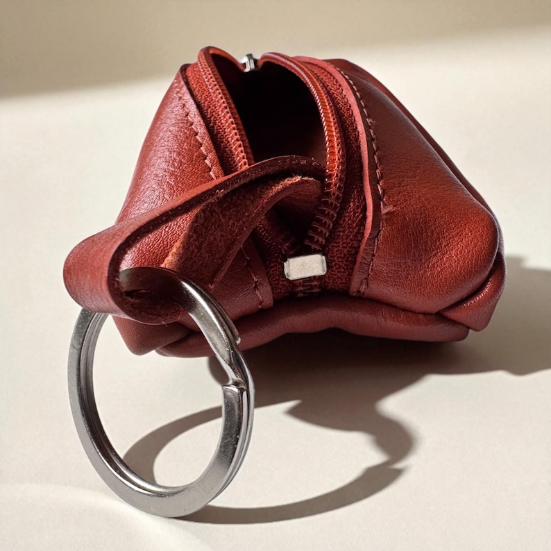 A small, tan leather coin purse with a silver key ring. The purse is open, showing the inside, and has a zip closure. The leather has a textured look, and the key ring is attached to a loop.