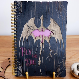 Puede incluir: Un cuaderno con espiral con un diseño de dragón cortado con láser en la portada. La portada es azul oscuro con un dragón dorado y las palabras "Fly or Die" en rosa. Un bolígrafo decorativo está en un soporte plateado.