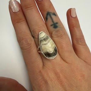 Puede incluir: Un anillo de plata con una piedra grande en forma de lágrima con un patrón único de remolinos crema, grises y negros. El anillo está engarzado en una banda de plata, mostrando la belleza natural de la piedra. El anillo se lleva en un dedo con uñas cuidadas.