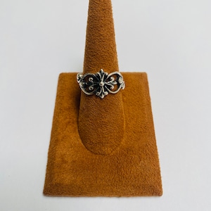 May include: A silver-colored ring with an ornate floral design. The ring features a central flower-like element with detailed petal patterns and symmetrical side accents. The ring is displayed on a brown ring holder against a white background.