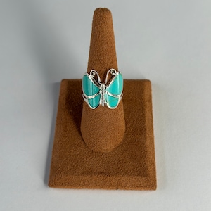 Puede incluir: Un anillo de mariposa turquesa con detalles plateados. El anillo se muestra en un soporte de anillo marrón.