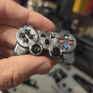 May include: A miniature silver-coloured games controller with black buttons and a directional pad. The controller has coloured buttons in red, green, and blue. The controller is held in a person's hand.