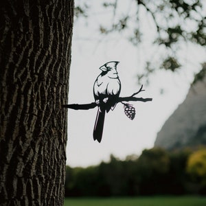 May include: A black metal silhouette of a cardinal perched on a branch with a pine cone. The silhouette is attached to a tree trunk.