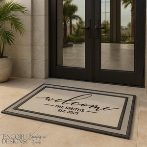 May include: Rectangular doormat with a light beige center and a dark gray border. The word "welcome" is written in a cursive font, with "THE SMITHS EST. 2025" below it. The mat is placed in front of a black double door.