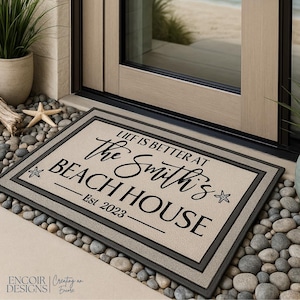 May include: Rectangular beige doormat with a black border and text that reads "LIFE IS BETTER AT the Smith's BEACH HOUSE Est 2023". The mat is placed on a bed of pebbles near a door.