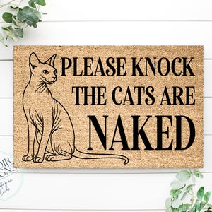 May include: A rectangular doormat with a Sphynx cat illustration and the text "PLEASE KNOCK THE CATS ARE NAKED" in black. The mat is a light brown color and is surrounded by greenery.