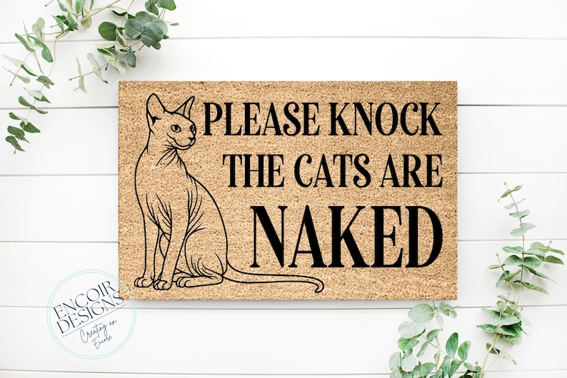 May include: A rectangular doormat with a Sphynx cat illustration and the text "PLEASE KNOCK THE CATS ARE NAKED" in black. The mat is a light brown color and is surrounded by greenery.