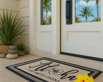 May include: A rectangular doormat with the word "Aloha" in cursive, a hibiscus flower, and "The Smith Ohana" in a serif font. The mat has a beige center, a gray border, and a black edge. Two yellow flowers are on the right.