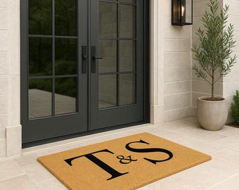 May include: A rectangular doormat with the letters "T & S" in black on a tan background. The mat is placed in front of a black double door. The image also shows a potted plant and a wall-mounted light.