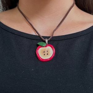 May include: A red apple pendant necklace with a green leaf and a tan center with four small holes. The pendant is on a brown cord necklace. The necklace is worn on a person wearing a black shirt.