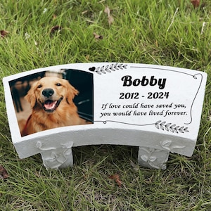 May include: A white stone bench with a photo of a golden retriever dog. The bench has the name "Bobby" and dates "2012-2024" with the text "If love could have saved you, you would have lived forever."
