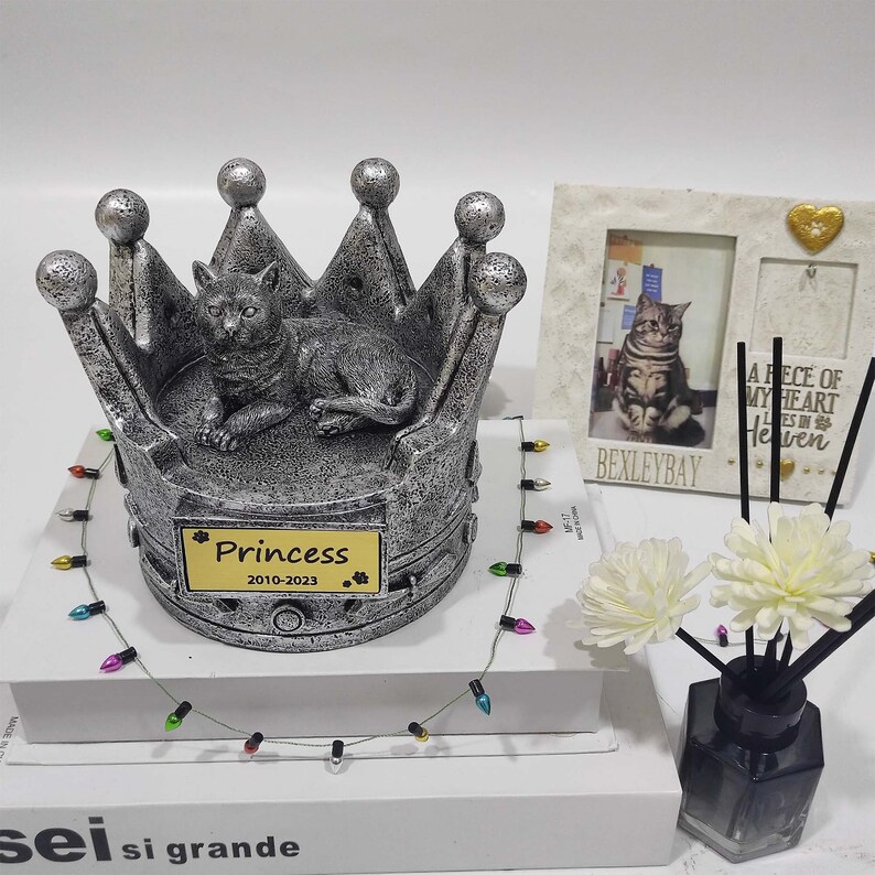 May include: A silver crown shaped pet urn with a cat figurine on top. The urn has a nameplate that reads "Princess 2010-2023".