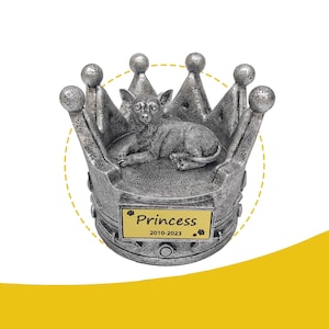 May include: A silver-colored cat figurine resting inside a crown. The crown has a detailed design with rounded tips. A gold plaque on the front reads "Princess 2010-2023". The cat is lying down, with its ears perked up.