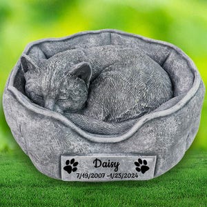 Puede incluir: Una cama conmemorativa para mascotas de concreto gris con una escultura de un gato durmiendo. La cama tiene una placa con el nombre "Daisy" y las fechas "7/19/2007 - 4/25/2024".