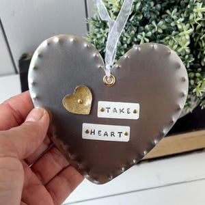 May include: A heart-shaped metal ornament with the words "TAKE HEART". The ornament is grey with a small gold heart attached. It has a white ribbon for hanging. The background includes greenery.