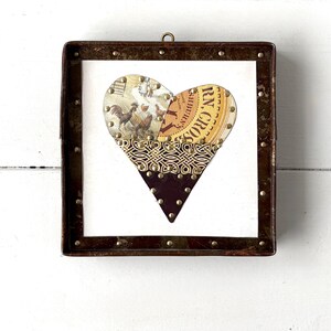 May include: A framed heart-shaped artwork with a rustic, vintage aesthetic. The heart is made from collaged paper, featuring rooster images and text, set against a white backdrop. The dark brown frame has brass-coloured studs.